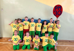 Dzieci stoją w grupie w strojach sportowych z transparentem naszego przedszkola w kształcie balona