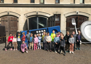 Dzieci z grupy 4 stoją przed muzeum, w tle widać duże rekwizyty talerz, widelec i telefon