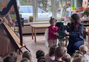 Pani gra na lirze, obok dziewczynka gra z pomocą na małej lirze