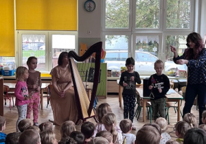Dzieci stoją na środku sali i grają na dzwonkach obok stoi lira