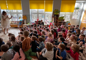 Dzieci siedzą na dywanie i śpiewają z prowadzącą piosenkę powitalną