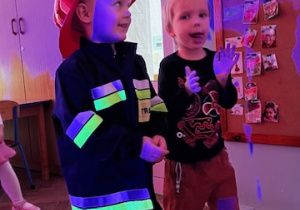 Chłopcy z grupy 1 stoją na podłodze, jeden w stroju strażaka