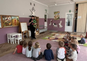 Dzieci z gr 1 siedzą na dywanie i słuchają jak pani prowadząca opowiada o wiankach wielkanocnych