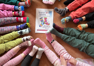 Dzieci siedzą na dywanie w kole, widać nogi i stopy w kolorowych skarpetkach