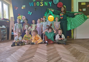 Zdjęcie grupowe grupy 3 z panią muzyczką, na tle kolorowej dekoracji wiosennej