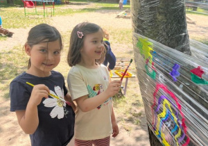 Dziewczynki z grupy 5 malują na folii zawieszonej pośród drzew w przedszkolnym ogrodzie