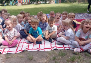 Dzieci z grupy 1 siedzą na kocu w przedszkolnym ogrodzie