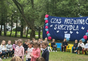 Dziewczynki z 5 grupy mówią wiersz stojąc w ogrodzie na tle dekoracji z napisem czas rozwinąć skrzydła