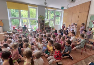 Dzieci siedzą na dywanie i słuchają koncertu. Z tyłu dzieczynki z 5 grupy tańczą